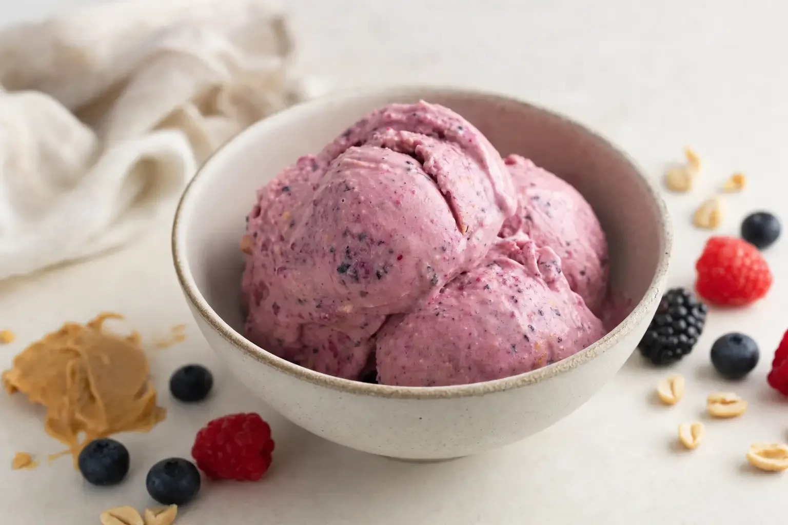 Bowl of scooped mixed berry ice cream surrounded by fresh berries, oats, and peanut butter